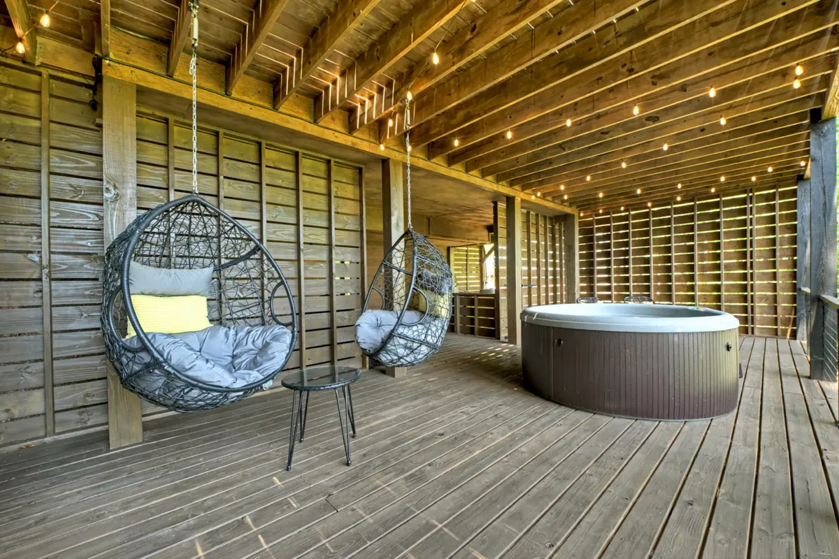 Hot tub on the lower covered deck beside two hanging egg chairs, lit by string lights along the timber ceiling.