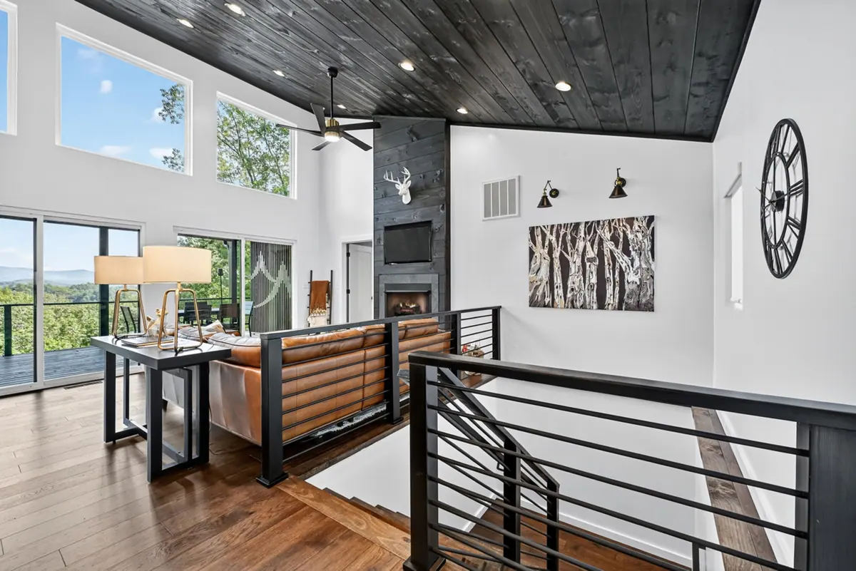 Open living room with a vaulted charred-timber ceiling, tan leather sofa and a stone fireplace, with the tree line visible through tall windows.