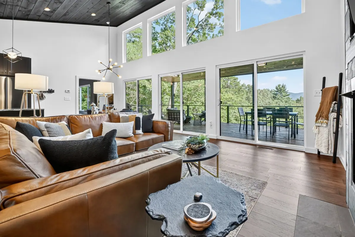 Tan leather sofa and armchairs arranged in front of the glass sliders, with the deck and forest beyond.