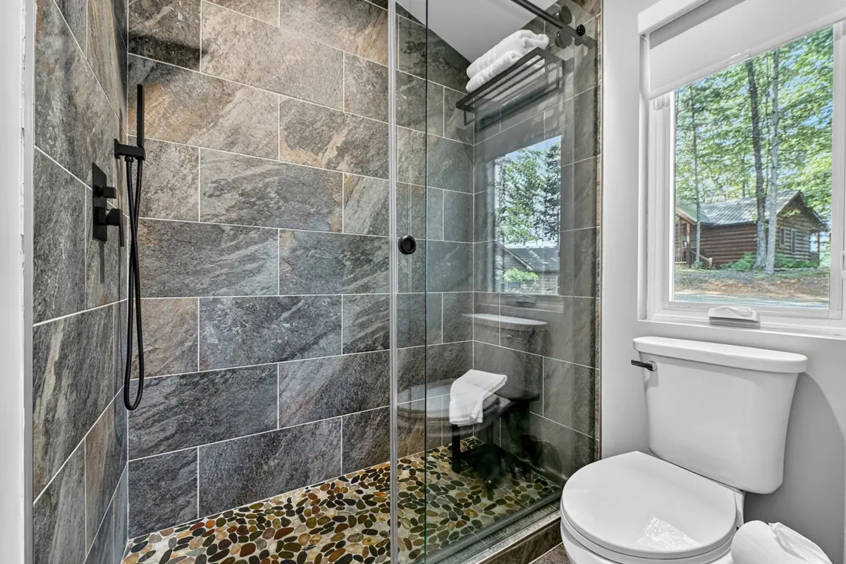 Walk-in shower tiled floor to ceiling in grey stone, with a pebbled floor and a window to the woods.