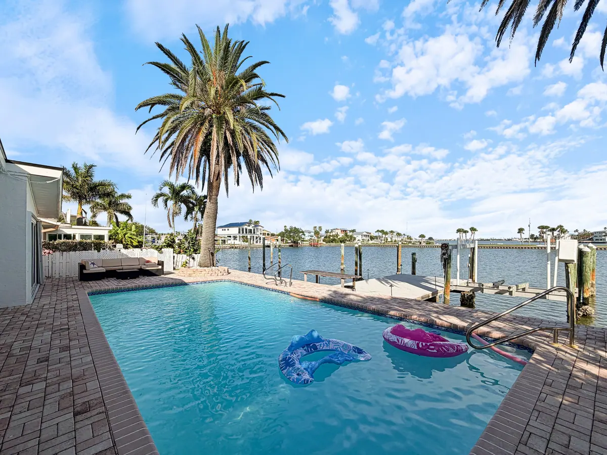 The pool in full sun with floats on the water, ringed by paver decking, palms and the dock beyond.