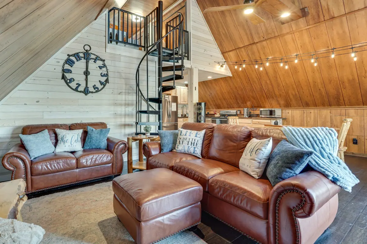 A-frame living room in warm pine with brown leather sofas, a spiral staircase and string lighting along the ceiling.