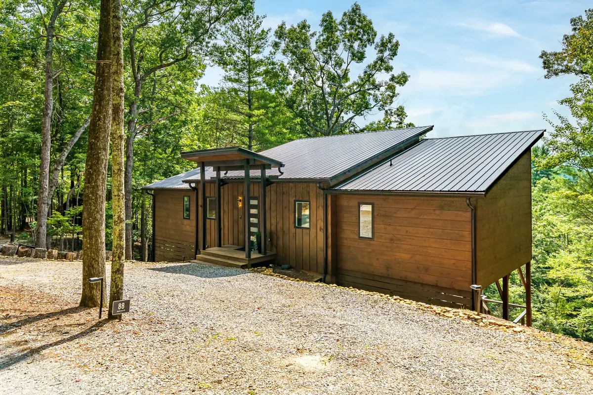 The cabin in daylight, timber-clad and raised among hardwoods at the end of a gravel drive.
