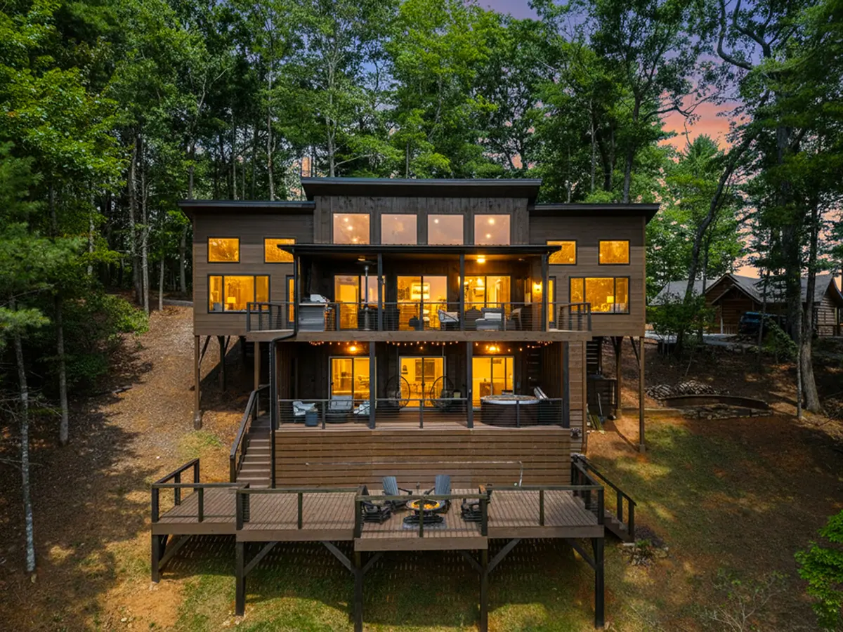 The cabin from the rear at dusk, three levels of glass and timber decking lit against the woods.