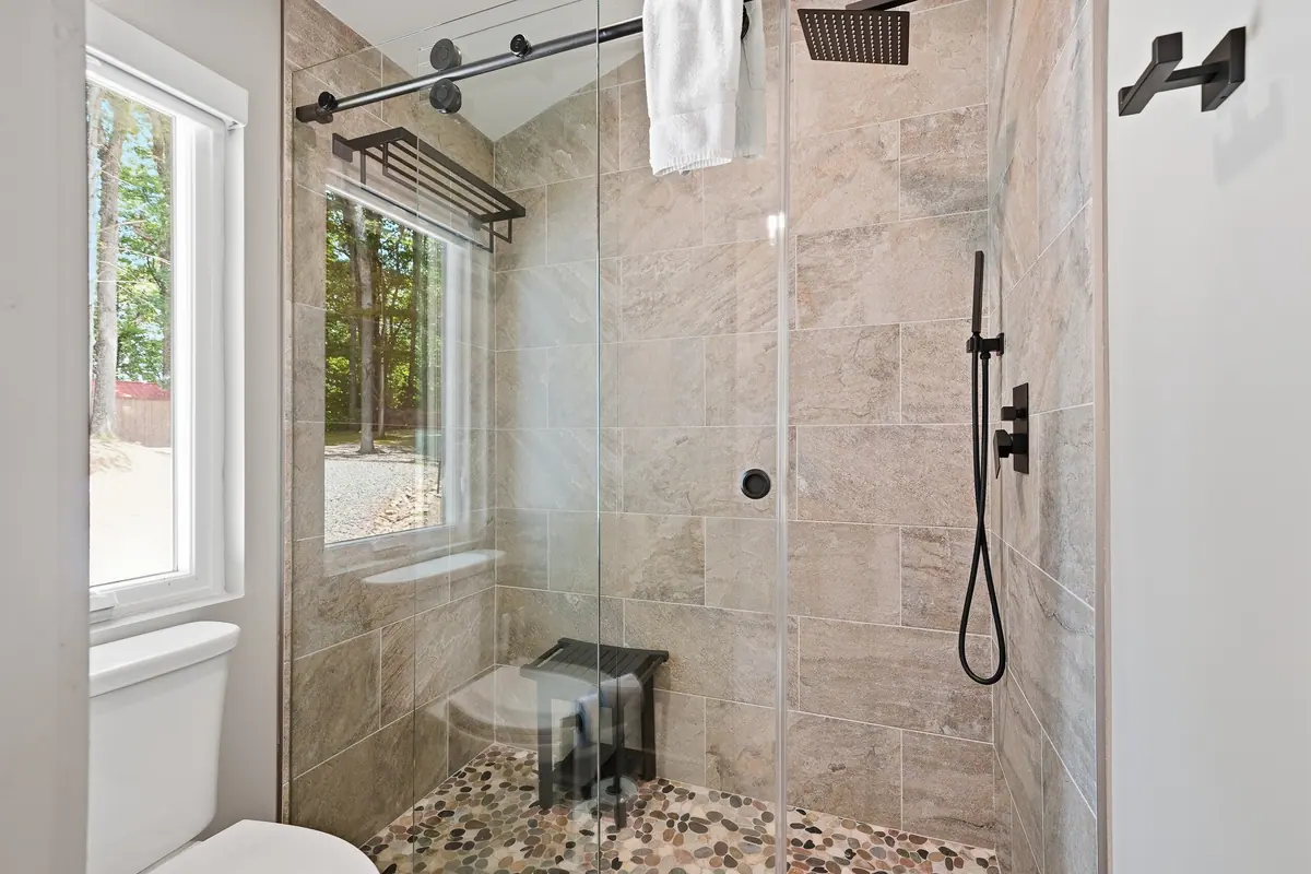 Walk-in shower tiled floor to ceiling in pale stone with a pebbled floor and a window to the trees.