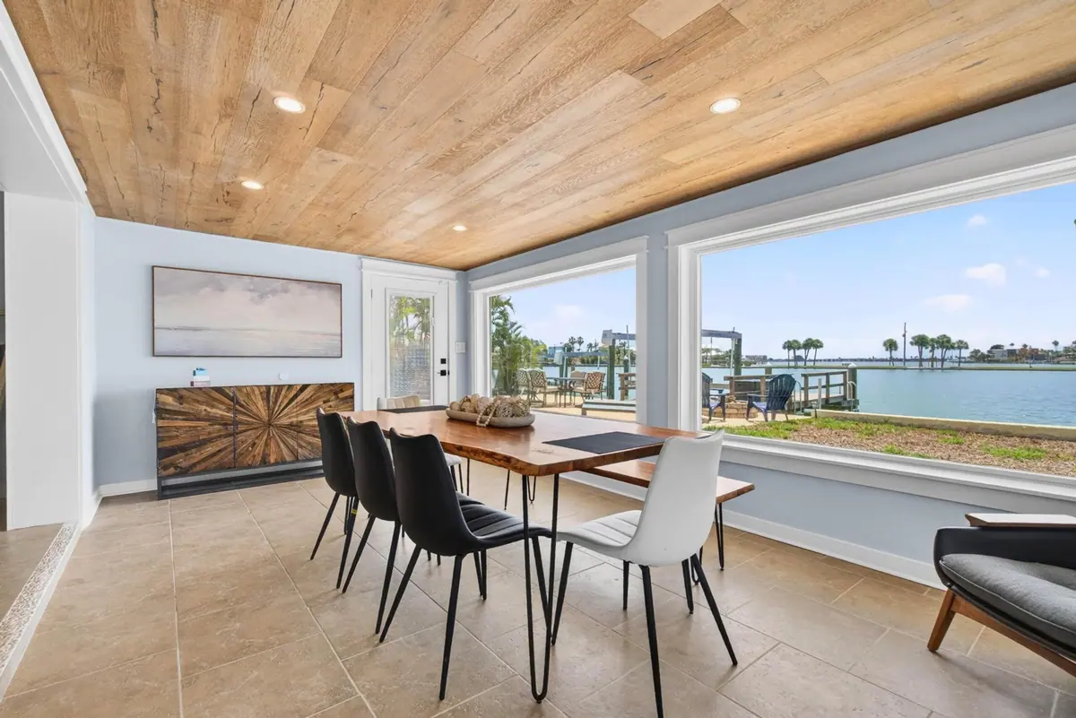 Dining table for six beneath a warm timber ceiling, set against a bank of windows looking straight out over the water.