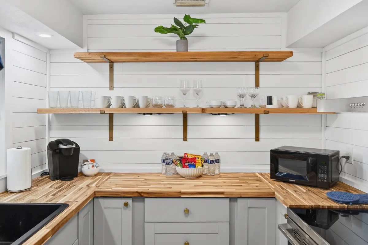 Kitchen with butcher-block counters, white cabinetry and open timber shelving stacked with everyday glassware.